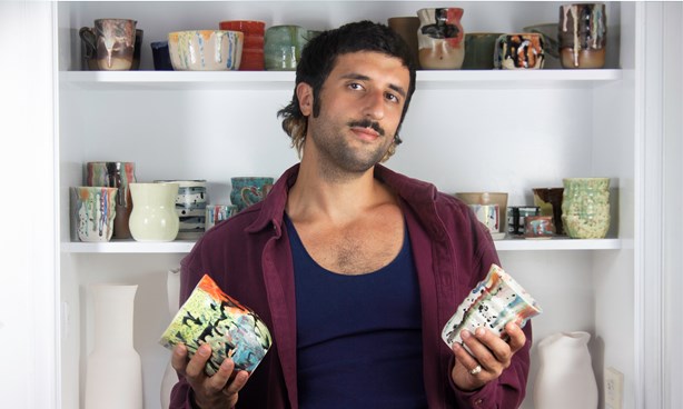 Headshot of Raphy Karanikos holding a ceramic vase in each hand with lots of ceramics on the shelves behind them