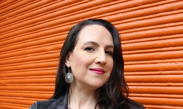 Headshot of Virginia Keft in front of an orange roller-door dressed elegantly in black