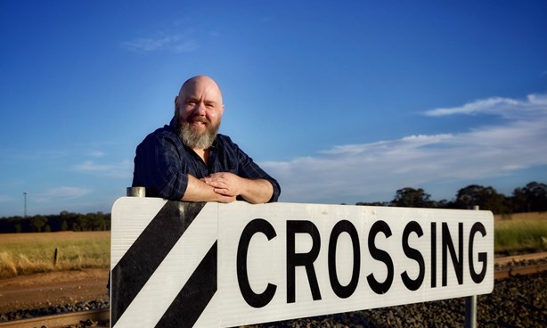 A bearded man standing behind a railway crossing sign