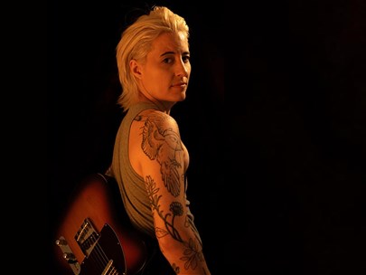 Female identifying person with lots of tattoos turned sideways to look at the camera against a black background.
