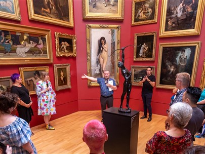A person presents a talk with a microphone to a crowd standing in front of a gallery wall at the National Gallery of Victoria, Melbourne.
