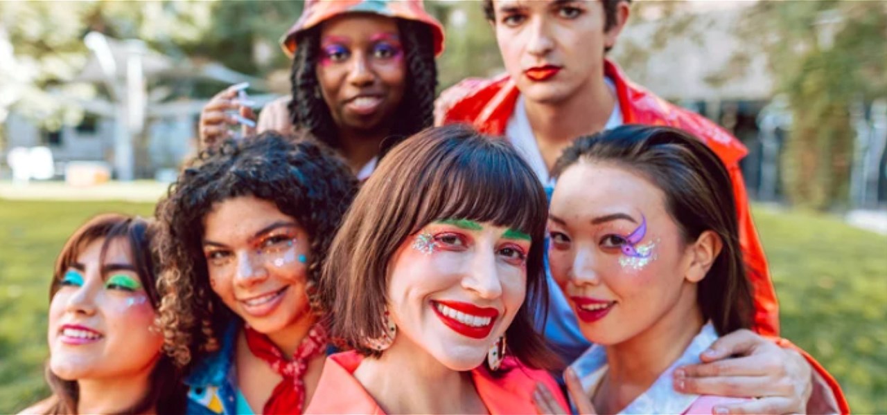 Six people in brightly coloured clothes and sparkly eye make-up are huddled together and smiling at the camera.