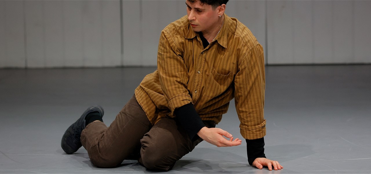 A person sits on the ground with his right arm gently crossing his body, and gazes towards the floor. He wears brown pants and a mustard shirt.