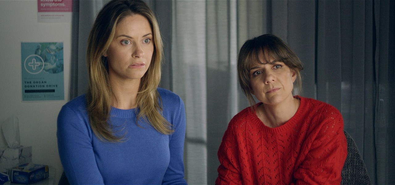 Two women sit facing ahead in a doctor's office, one wearing a red sweater and the other wearing a blue sweater, both looking worried.