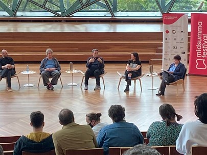 6 people sit on chairs in a panel discussion in a room with glass walls and wooden floor