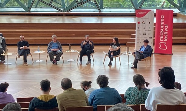 6 people sit on chairs in a panel discussion in a room with glass walls and wooden floor