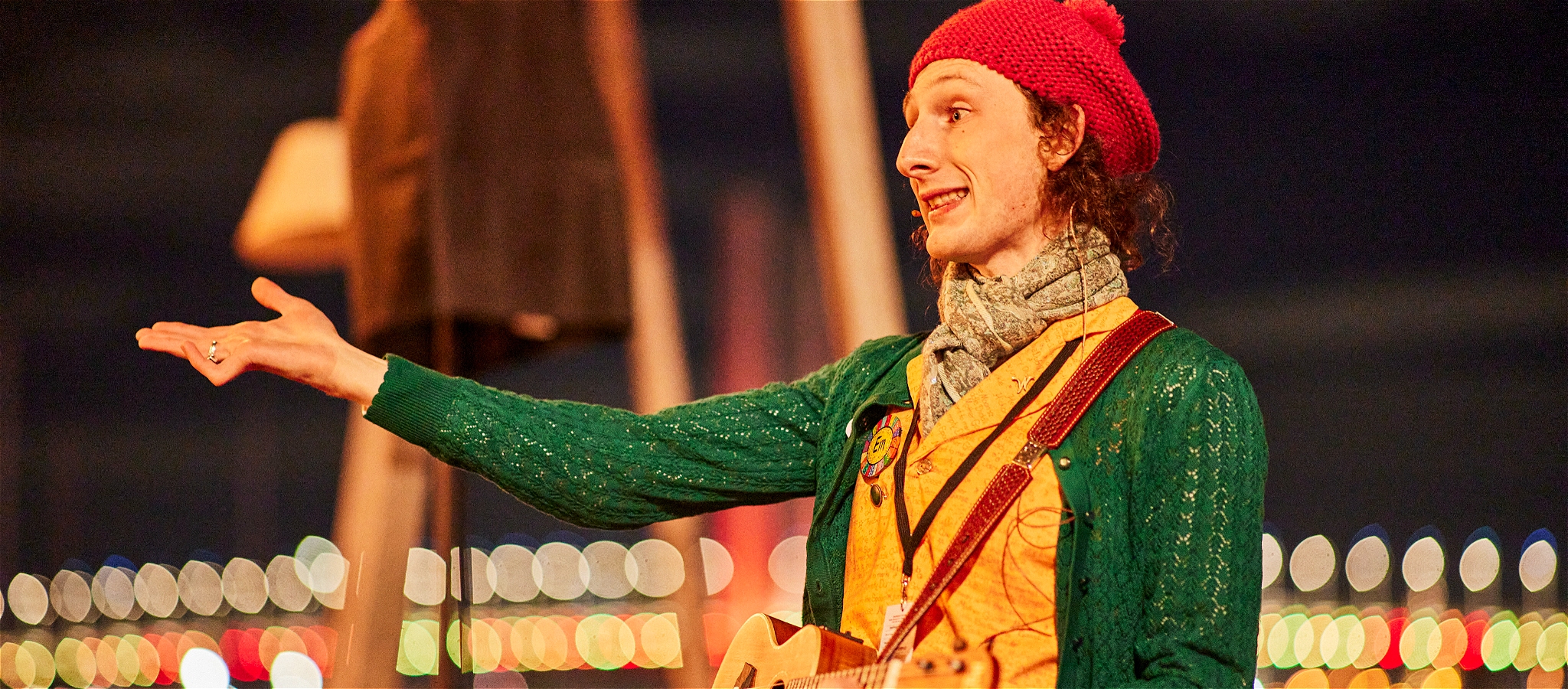 A person wearing a green cardigan, yellow vest and pink hat holding their hand out.