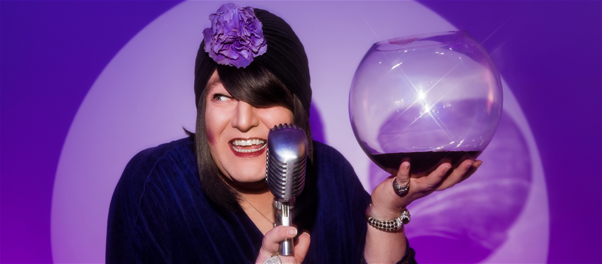 Tina Del Twist smiling behind an old microphone holding a fishbowl of red wine against purple bubble shaped background