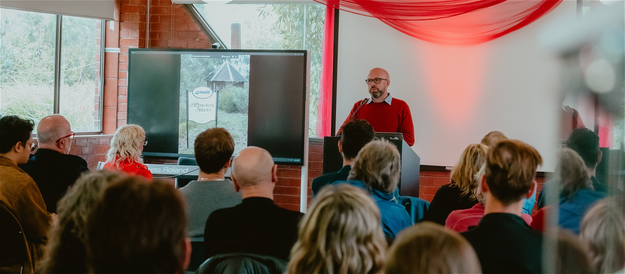 A man stands in front of a podium speaking to a crowd of people seated in front. A screen and digital slideshow is being presented next to the speaker
