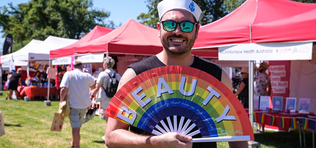 Person wearing sailor hat holding an opened rainbow fan with text "BEAUTY", standing in front of Midsumma Carnival stalls
