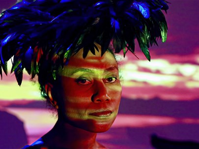 A Pacific Islander person wearing a feather headdress with a landscape at dusk and their shadow behind them