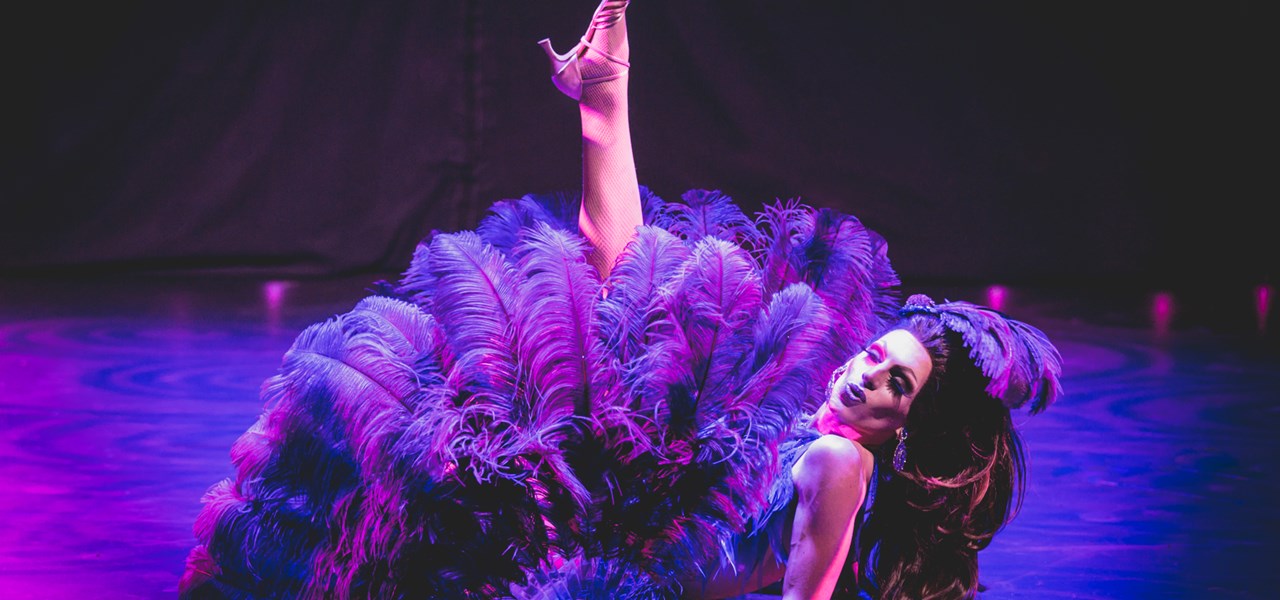 Female-identifying person dressed in a large feather dress, lying on stage, one leg in air. Mauve lighting
