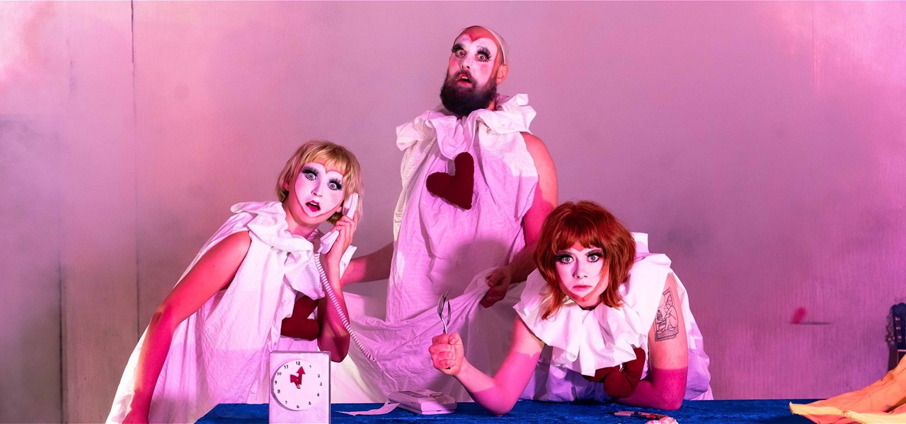Three people dressed in white nightdresses with puffy collars are gathered around a table. They are painted like mimes and hold odd expressions.