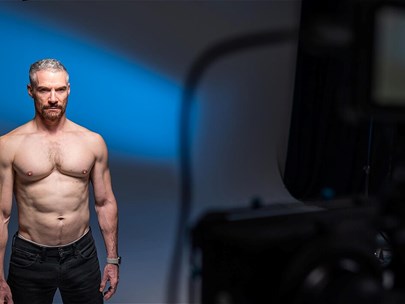 A muscular, shirtless man wearing jeans poses in front of a camera in a photography studio.