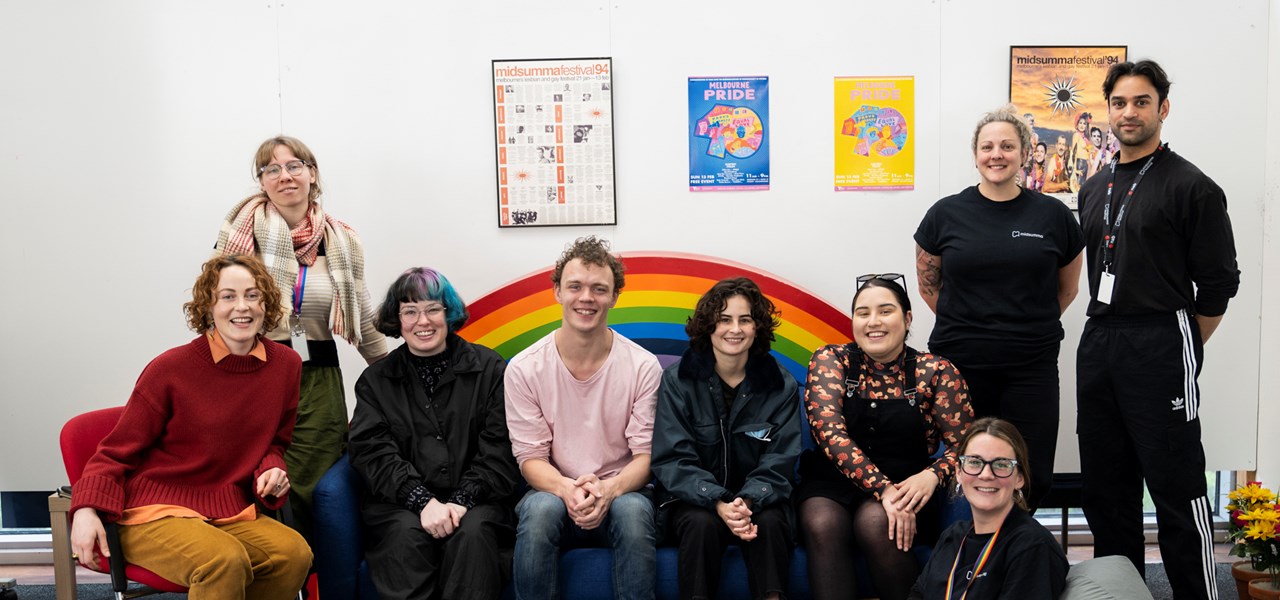 People seated or standing around a couch, posing for the camera, with some midsumma guide posters behind them