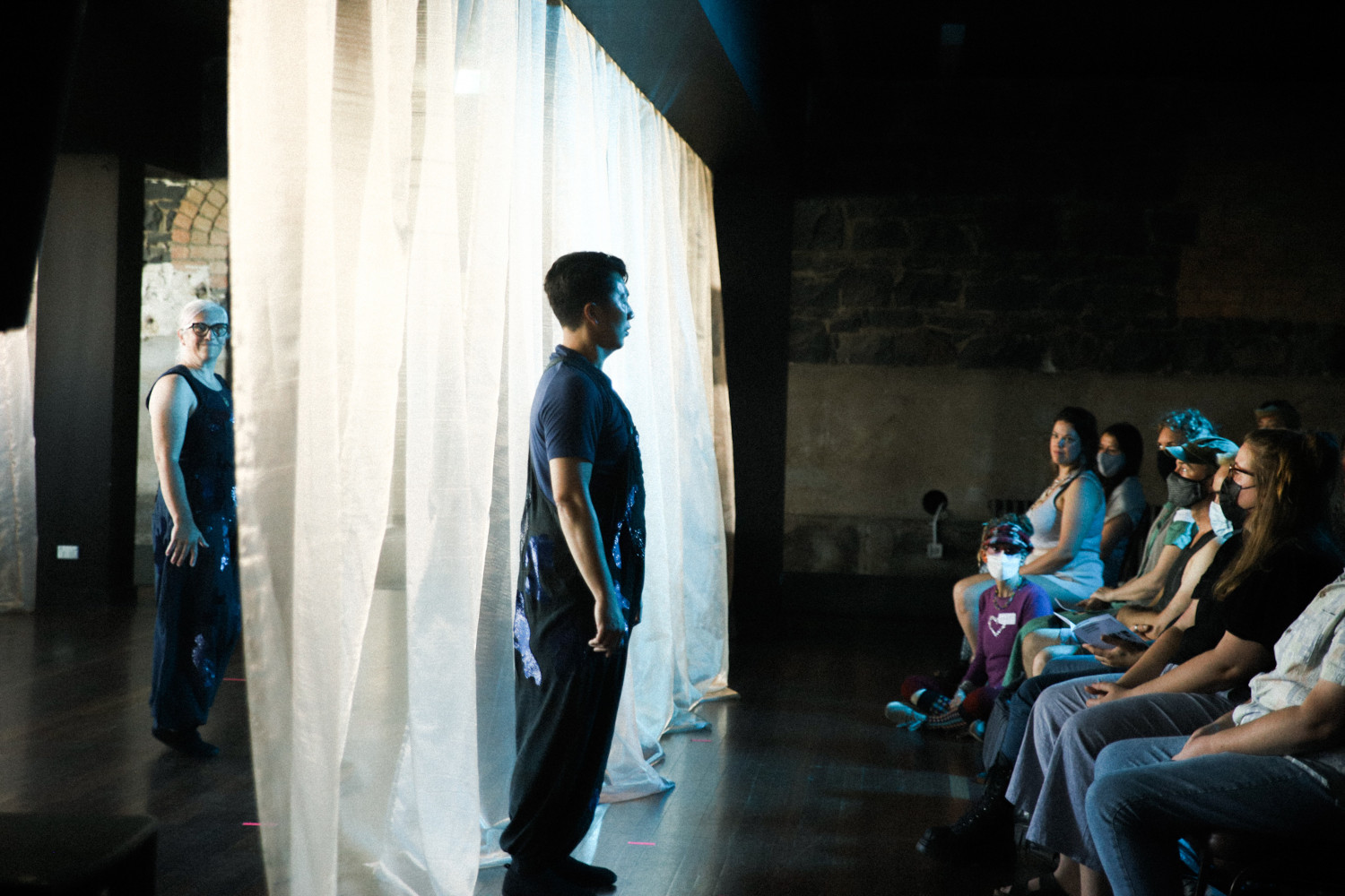 A person standing in front of a curtain facing an audience, with another person hidden behind the curtain