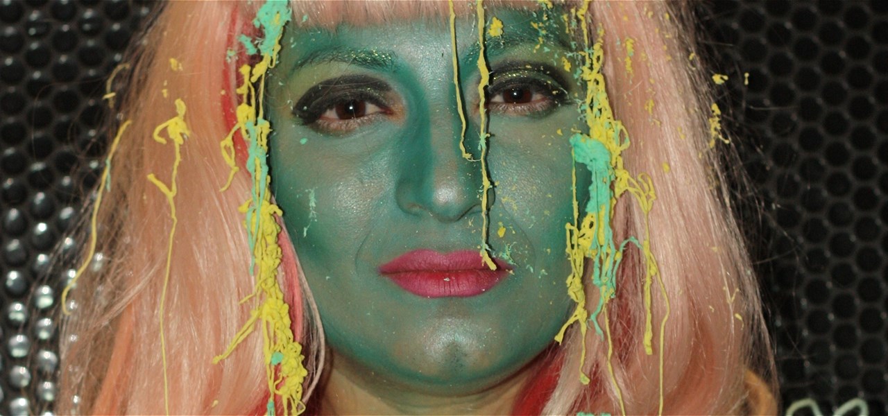 Head of a person whose face is painted green with red lipstick and blonde wig looking musingly at the camera