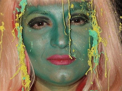 Head of a person whose face is painted green with red lipstick and blonde wig looking musingly at the camera