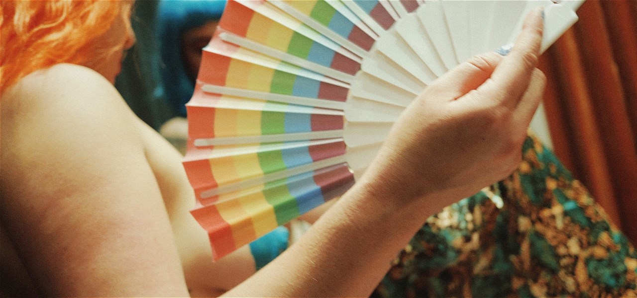 A nude person with orange hair holding a rainbow fan.