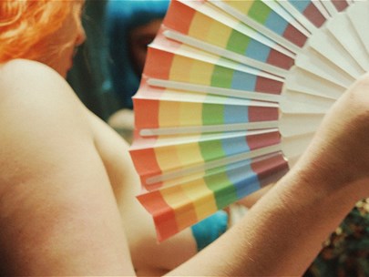 A nude person with orange hair holding a rainbow fan.