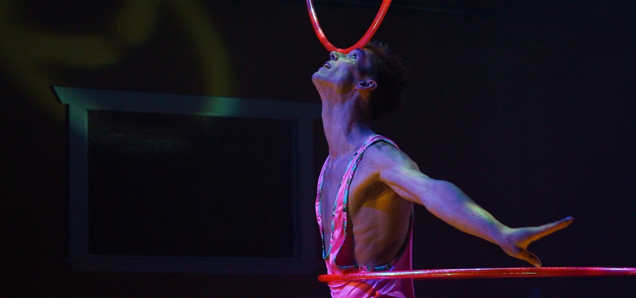 A person balancing a hula hoop on his head while hula hooping around their waist. They are wearing a pink leotard