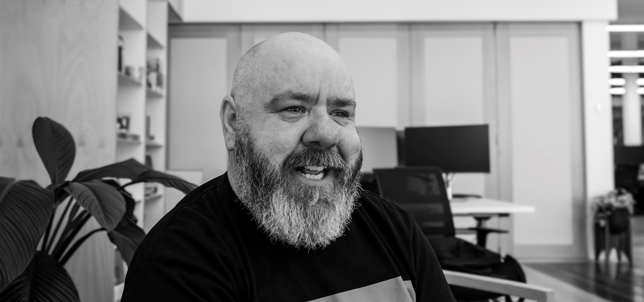 Black and white image of Ben smiling in a room with a plant and sliding doors behind him