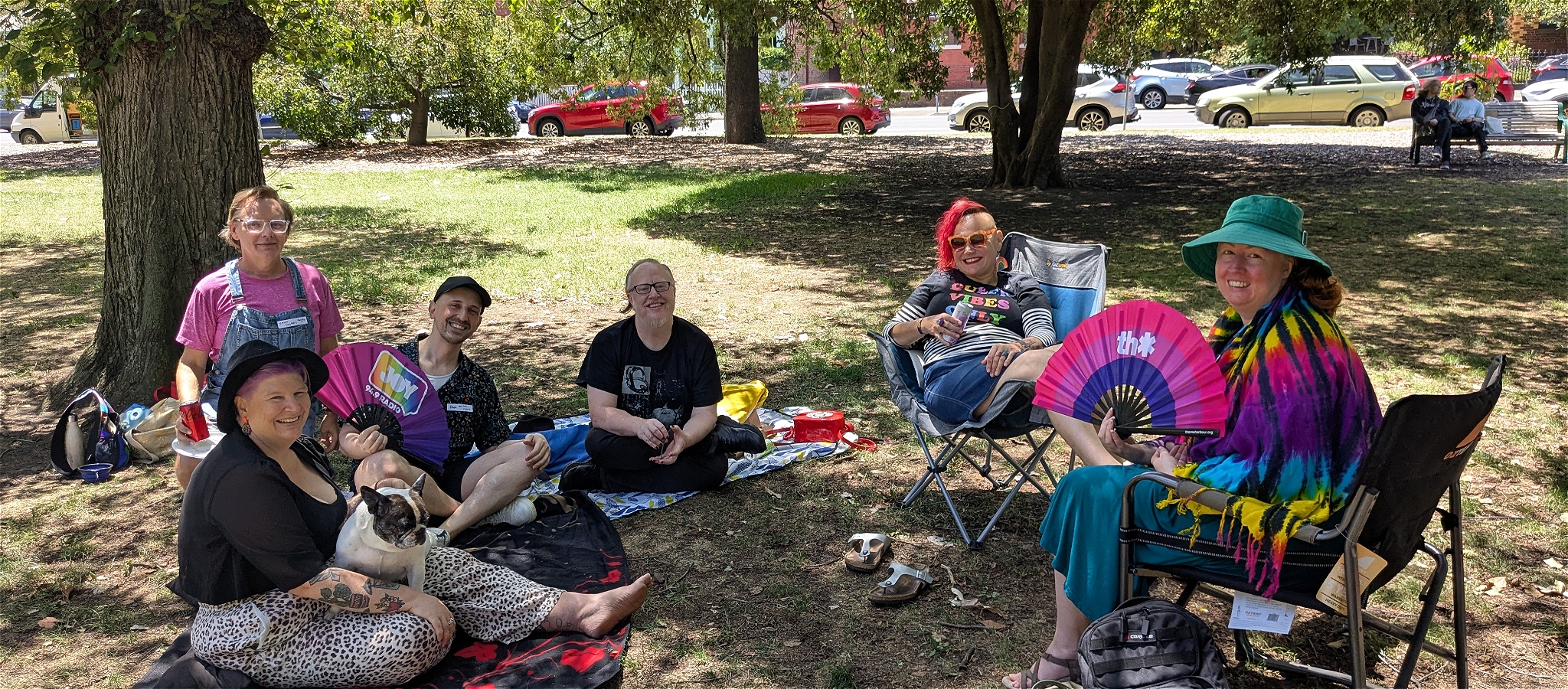 A group of smiling people with brightly coloured hair and clothes, sitting around a picnic rug with a bisexual flag visible in the foreground.