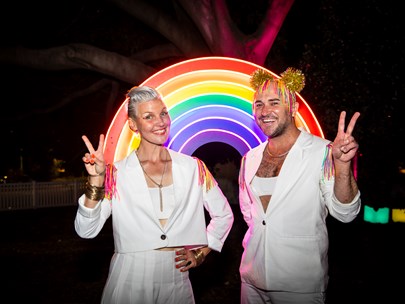 Bec Reid and Tristan Meecham happily giving a victory sign while standing in front of a rainbow arch