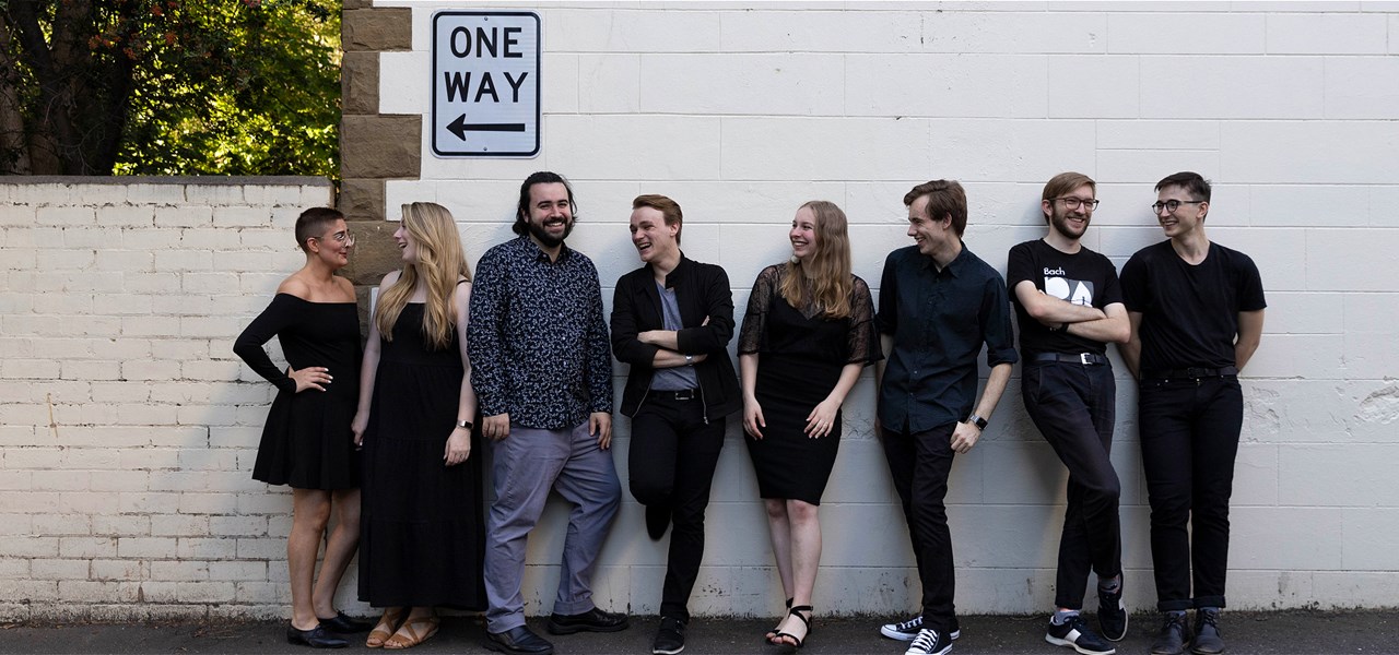 Eight singers wearing black stand in front of a white wall smiling at each other