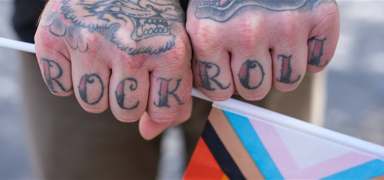 Two fists holding a Pride Flag with 'Rock Roll' tattooed across the knuckles.