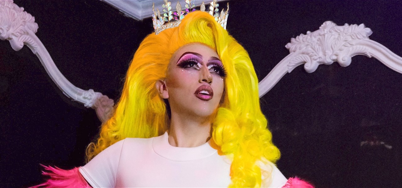 Drag Queen in a light pink t-shirt with bright pink feathers on the sleeves and a bright yellow wig and crown