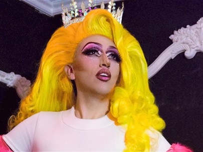 Drag Queen in a light pink t-shirt with bright pink feathers on the sleeves and a bright yellow wig and crown