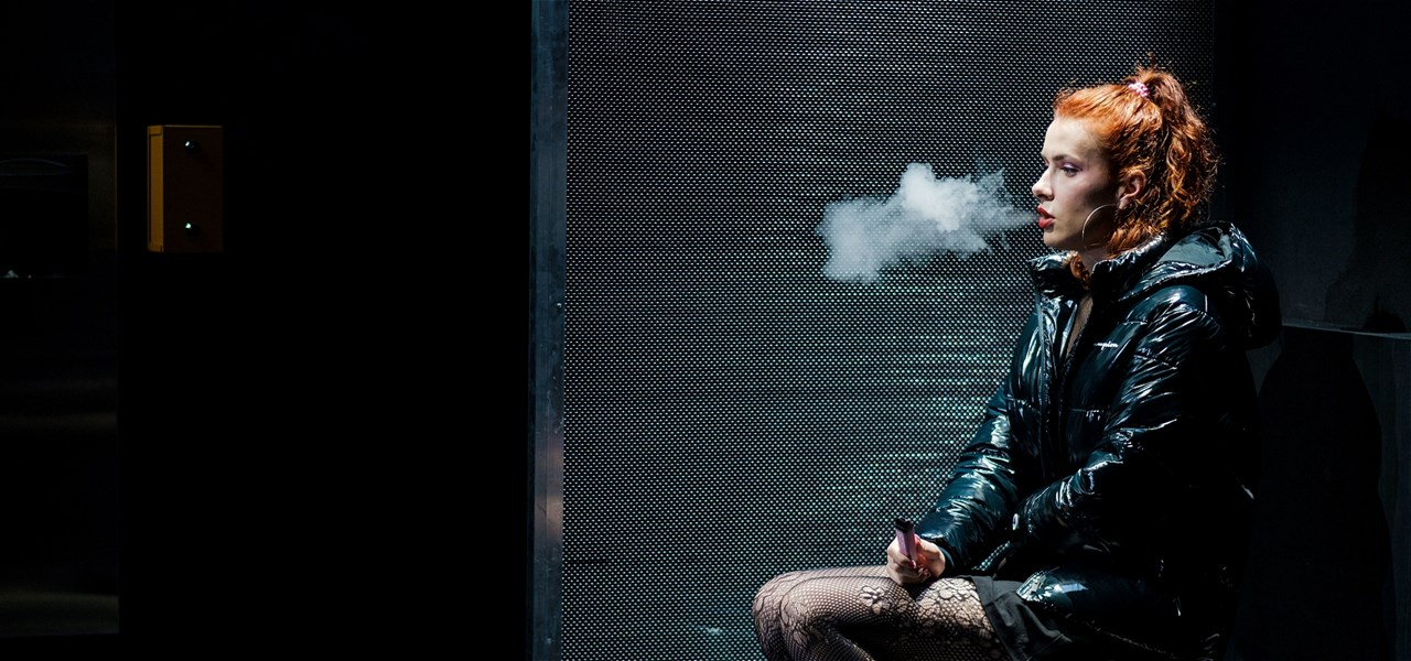 Person with orange hair and black jacket sitting down vaping.