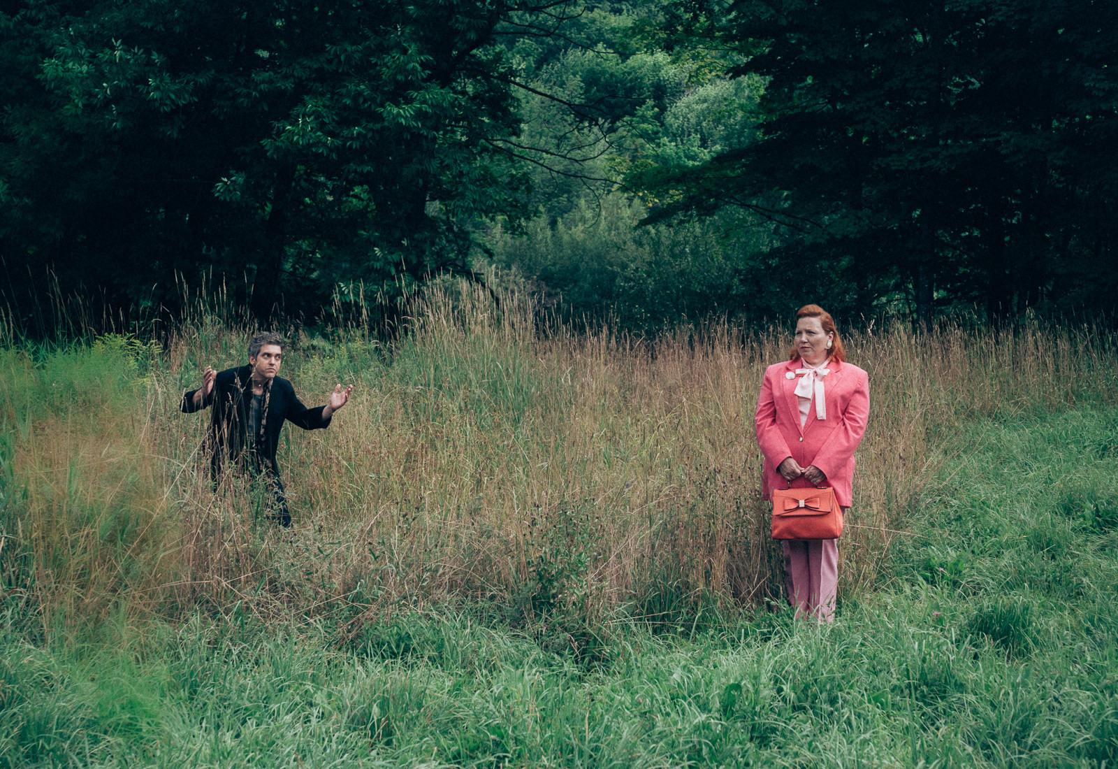 Two people in a grassland setting, one a well dressed woman in puce, the other a man holding their hands in the air
