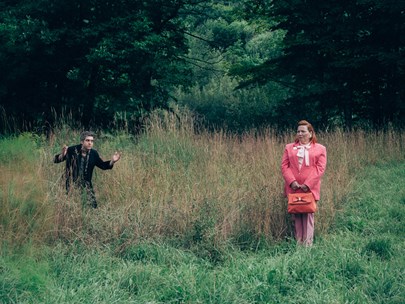 Two people in a grassland setting, one a well dressed woman in puce, the other a man holding their hands in the air