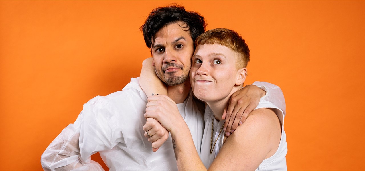 Two individuals standing in front of an orange background holding each other.