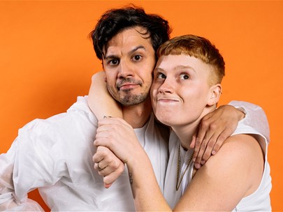 Two individuals standing in front of an orange background holding each other.