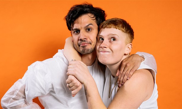 Two individuals standing in front of an orange background holding each other.