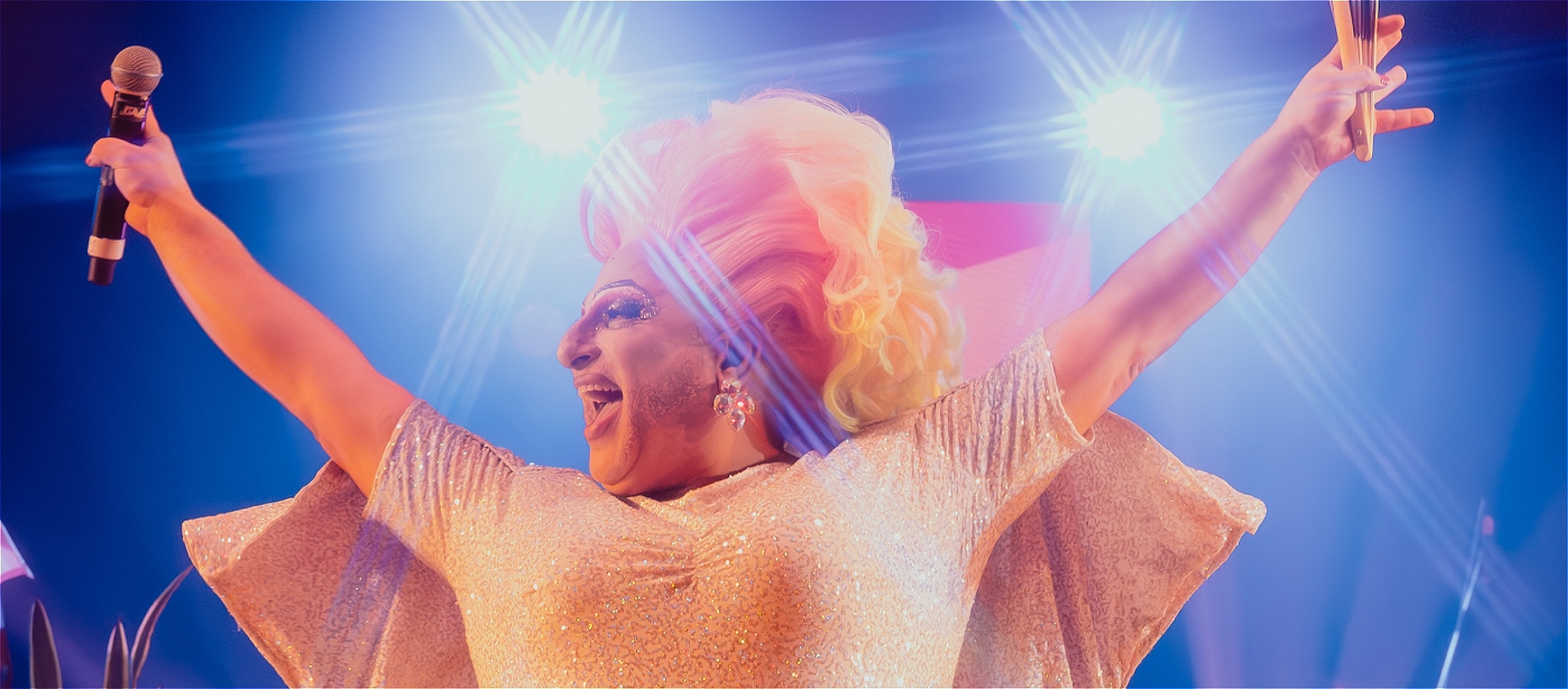 Drag star Frock Hudson holds a microphone and fan high, in a glittering sequinned gown under stage lights.