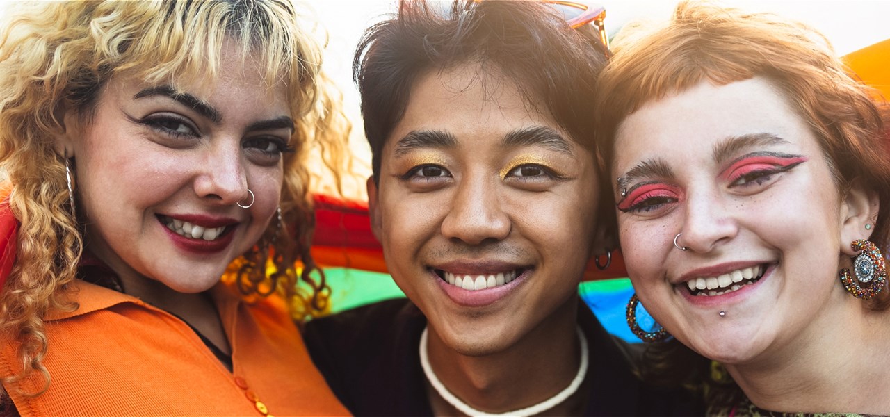 Three friends smiling in front of a rainbow flag