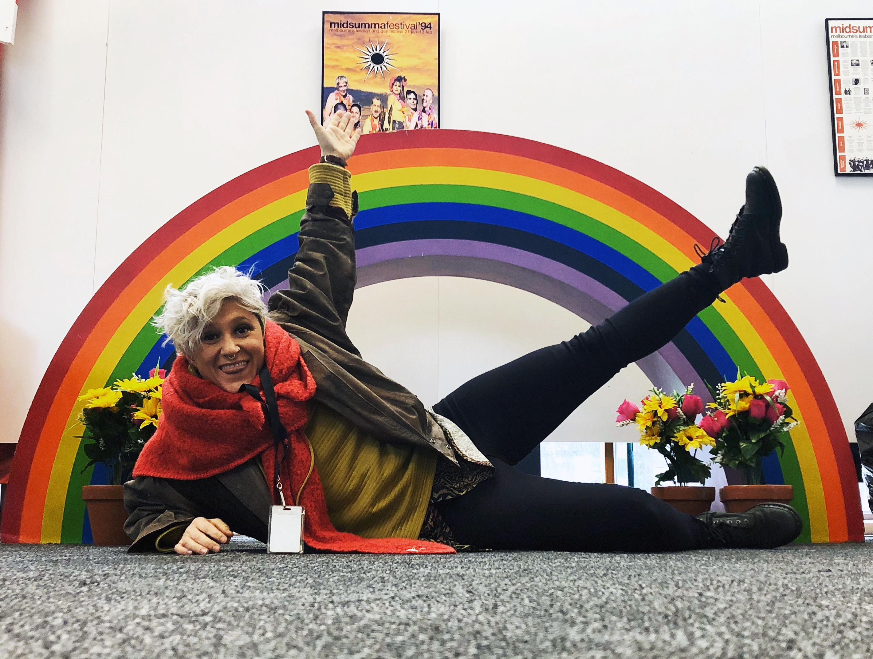 Sasha lying on the floor, one arm and one leg raised, in front of a rainbow arch