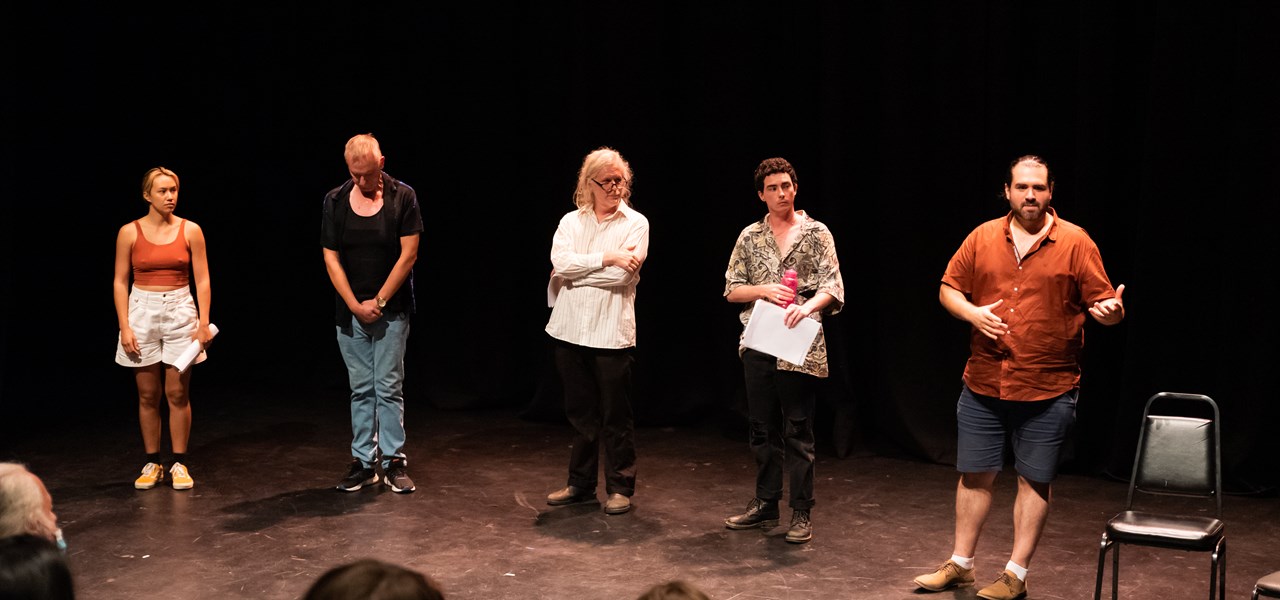 A number of actors standing on a stage, several with scripts in their hands