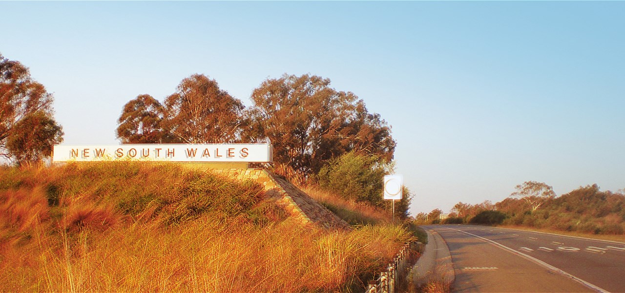 A border sign reading "NEW SOUTH WALES" beside the highway. There's native grasses in the foreground and eucalyptus trees in the background.
