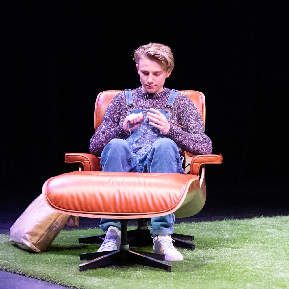 Man dressed in overalls and knitted jumper sitting on leather chair, knitting