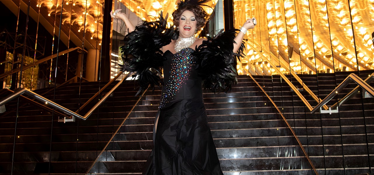 Dolly stands on the steps of a theatre. Lights and mirrors surround her. She is laughing with her hands in the air and wearing a black gown.