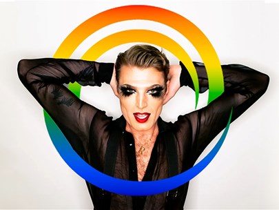 Reuben Kaye stands in a black open neck sheer shirt with heavy eye makeup and red lipstick with his arms behind his head inside a rainbow swirl.