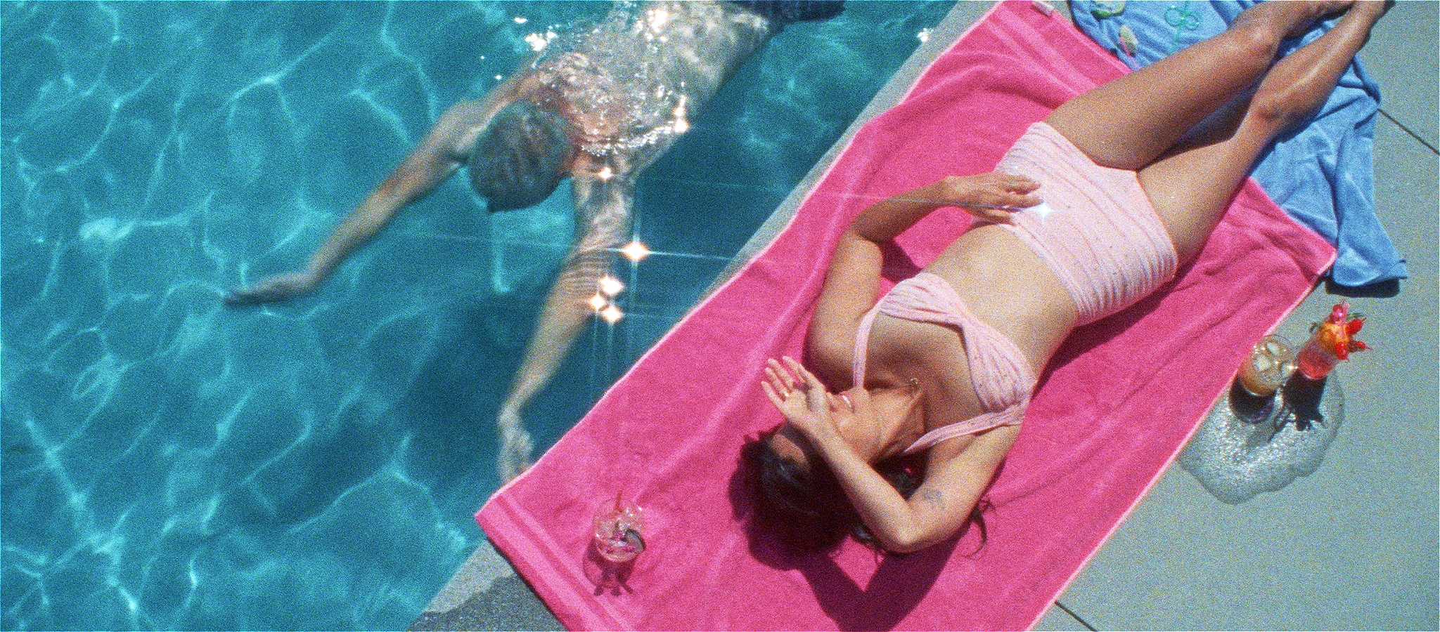 a person swimming underwater in a pool next to someone in a pink bikini laying poolside on a pink towel with two cocktails next to them