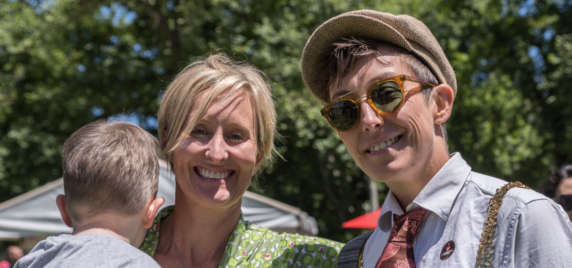 Two adults smiling and holding a baby. Outdoors on a sunny day. One has blond hair and the other is wearing a vintage looking newspaper boy cap, suspenders and a tie.