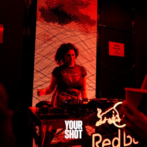 Stevue Ellis spinning the decks. Lighting is dark with red tones.