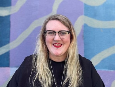 Rhen smiles into the camera while standing in front of a multicoloured background. They have long blonde hair and are wearing glasses and red lipstick.